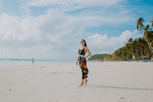 Full body of slim barefoot female tourist wearing black summer outfit and sunglasses standing hand on waist on white sandy beach near sea under cloudy blue sky