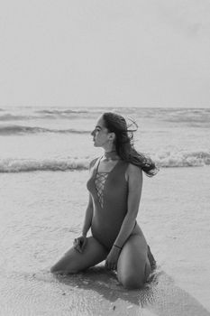 Black and white portrait of a woman kneeling on the beach, sea waves in the background.