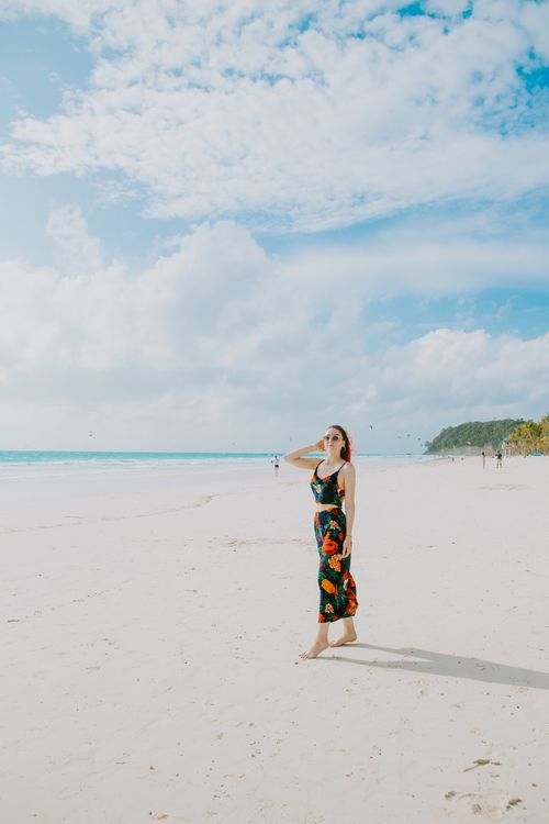 Free Woman in a floral dress walking on a sunlit beach, enjoying a peaceful day by the sea. Stock Photo