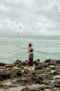 Woman in a floral dress standing on a rocky beach, watching colorful windsurf sails on a cloudy day.