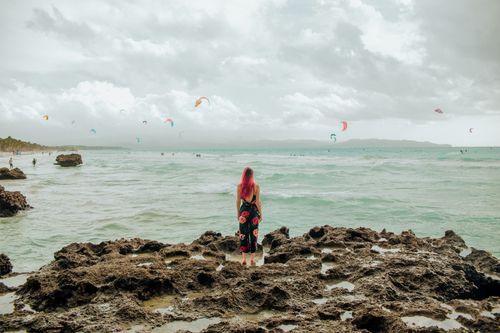Free A woman with pink hair stands on a rocky seashore, overlooking the ocean under a cloudy sky with kite surfers. Stock Photo