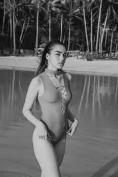Stylish woman in swimsuit on tropical beach with palm trees in black and white photo.