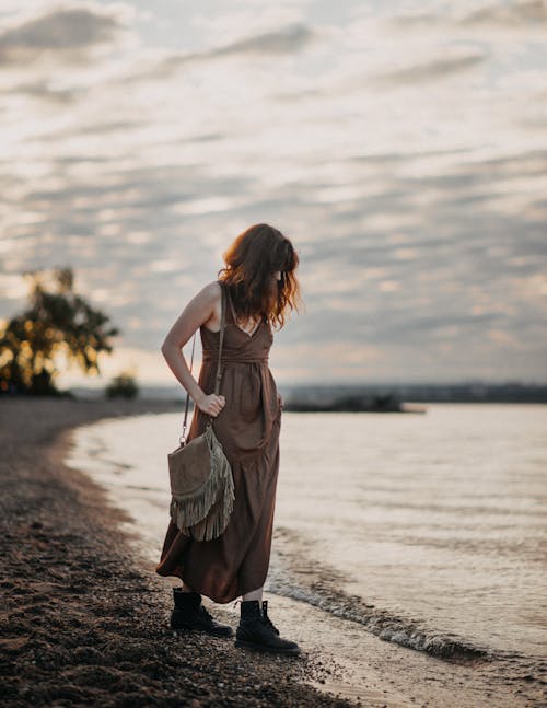 Full body of lady in brown dress standing on beach near waving sea and looking down in twilight