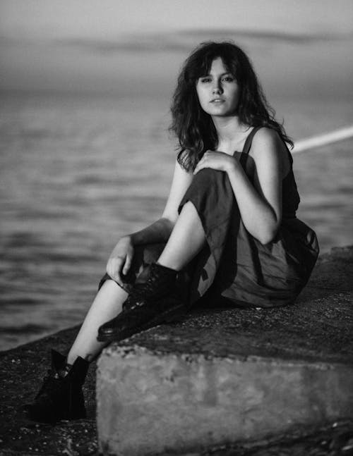 Black and white of female sitting on stone near sea and looking at camera dreamily