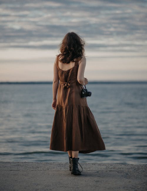 Back view of unrecognizable female in stylish dress and boots with photo camera standing near blurred rippled sea in dusk