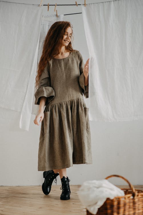 Full body of charming young female in stylish dress standing near wicker basket and white fabric hanging on rope in apartment