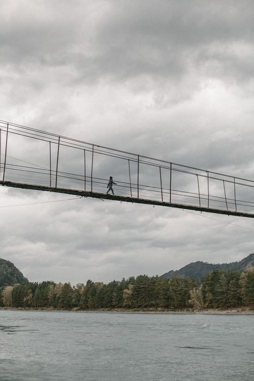 Free A person crossing a suspension bridge against a cloudy sky with scenic river and forest views. Stock Photo