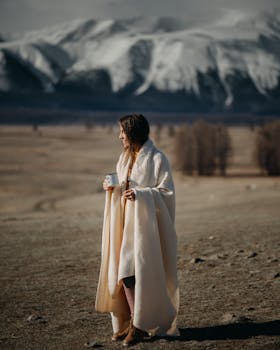 Young woman wrapped in a blanket enjoying coffee against a breathtaking mountain backdrop in autumn.