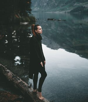 Side view full body of barefoot female in black clothes standing on log near calm lake in mountainous area and looking away