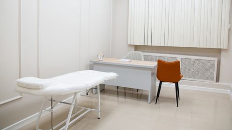 A minimalist medical examination room featuring a desk, chairs, and an examination bed in a sterile, neutral setting.