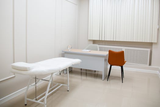 A minimalist medical examination room featuring a desk, chairs, and an examination bed in a sterile, neutral setting.