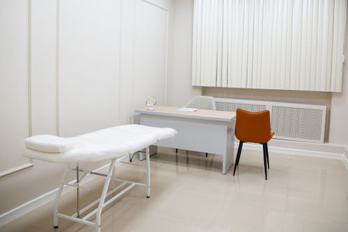 Free A minimalist medical examination room featuring a desk, chairs, and an examination bed in a sterile, neutral setting. Stock Photo