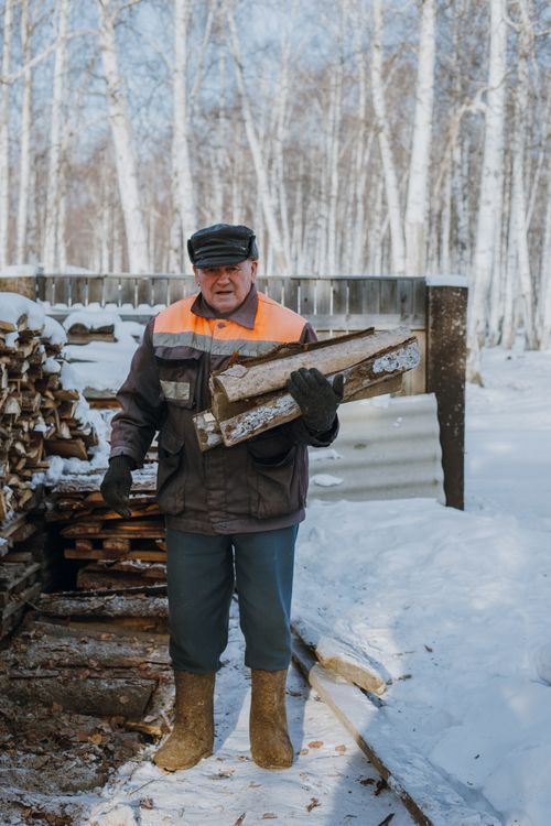 Free An elderly man dressed warmly carries firewood in a snowy winter forest scene. Stock Photo