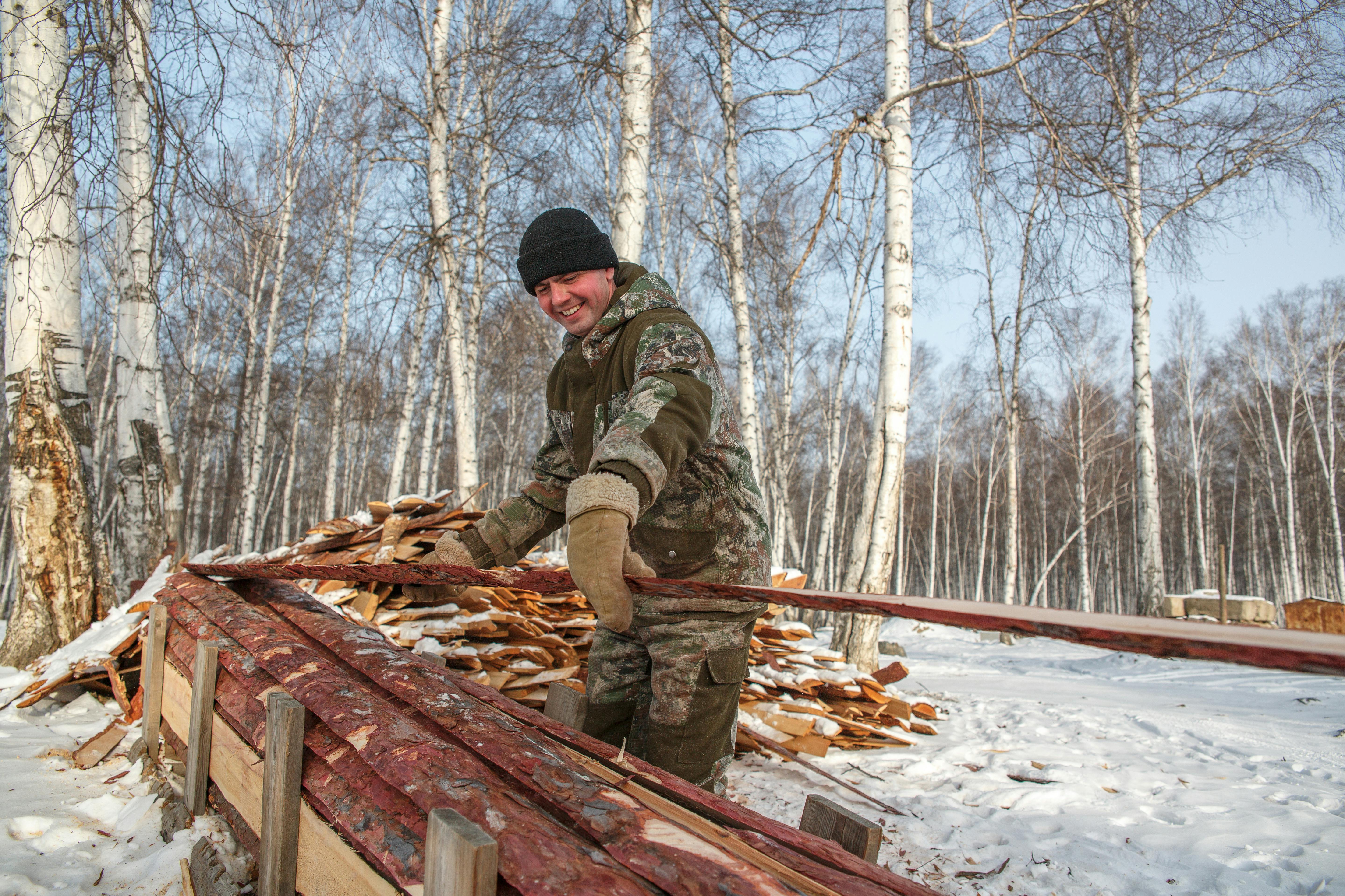 Smiling man handling wood in a snow-covered forest setting during winter.