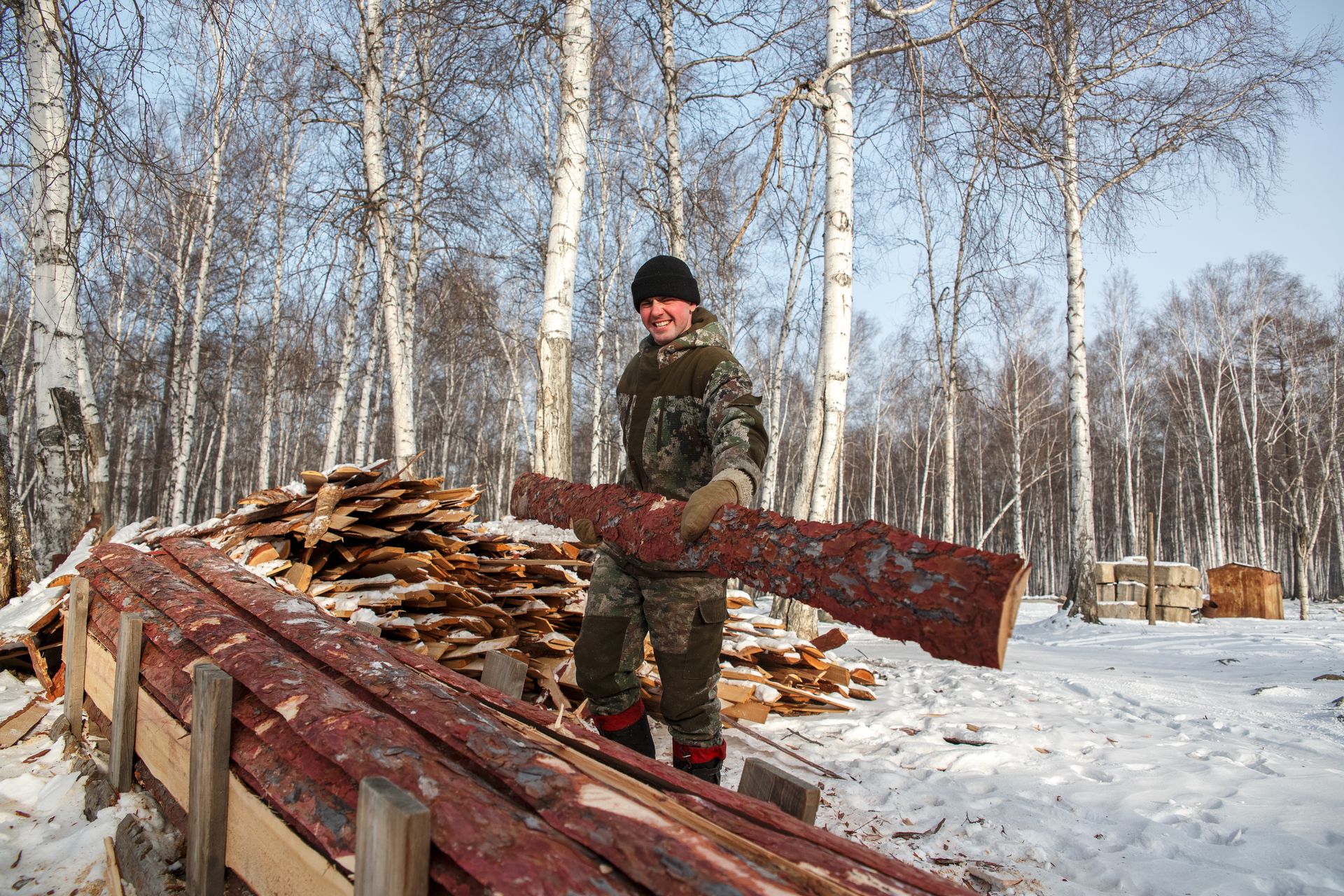 A man stacks firewood in a snowy forest during winter, dressed warmly for the cold.