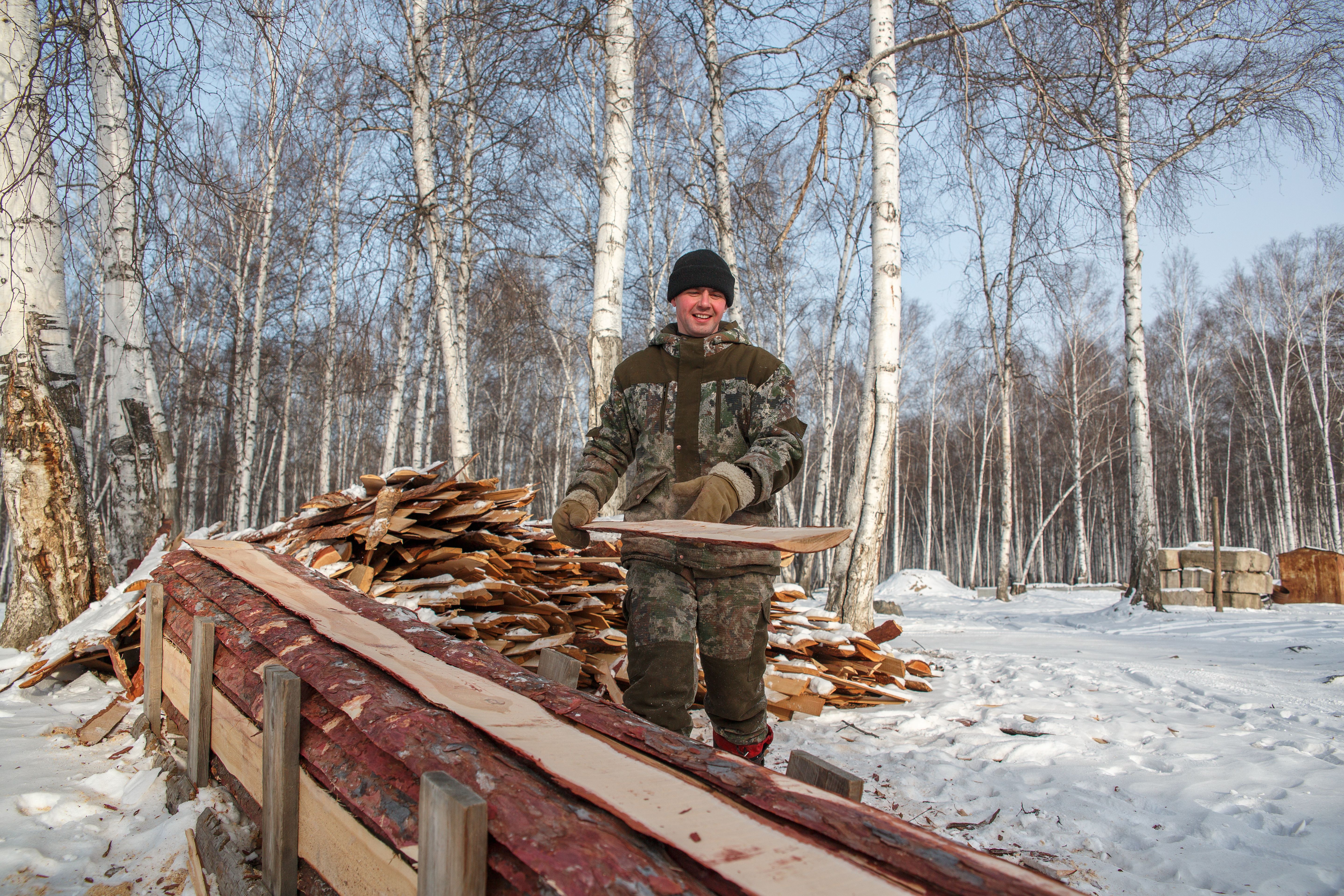 A man smiling while stacking wood in a snowy birch forest during winter.