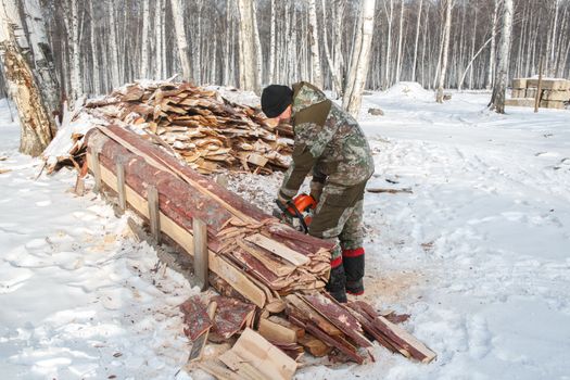 A man in camouflage gear cuts firewood with a saw amidst a snowy forest setting, surrounded by trees and wood stacks.