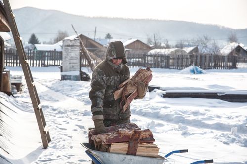 Free A man in winter clothing handles firewood in a snowy village landscape. Stock Photo