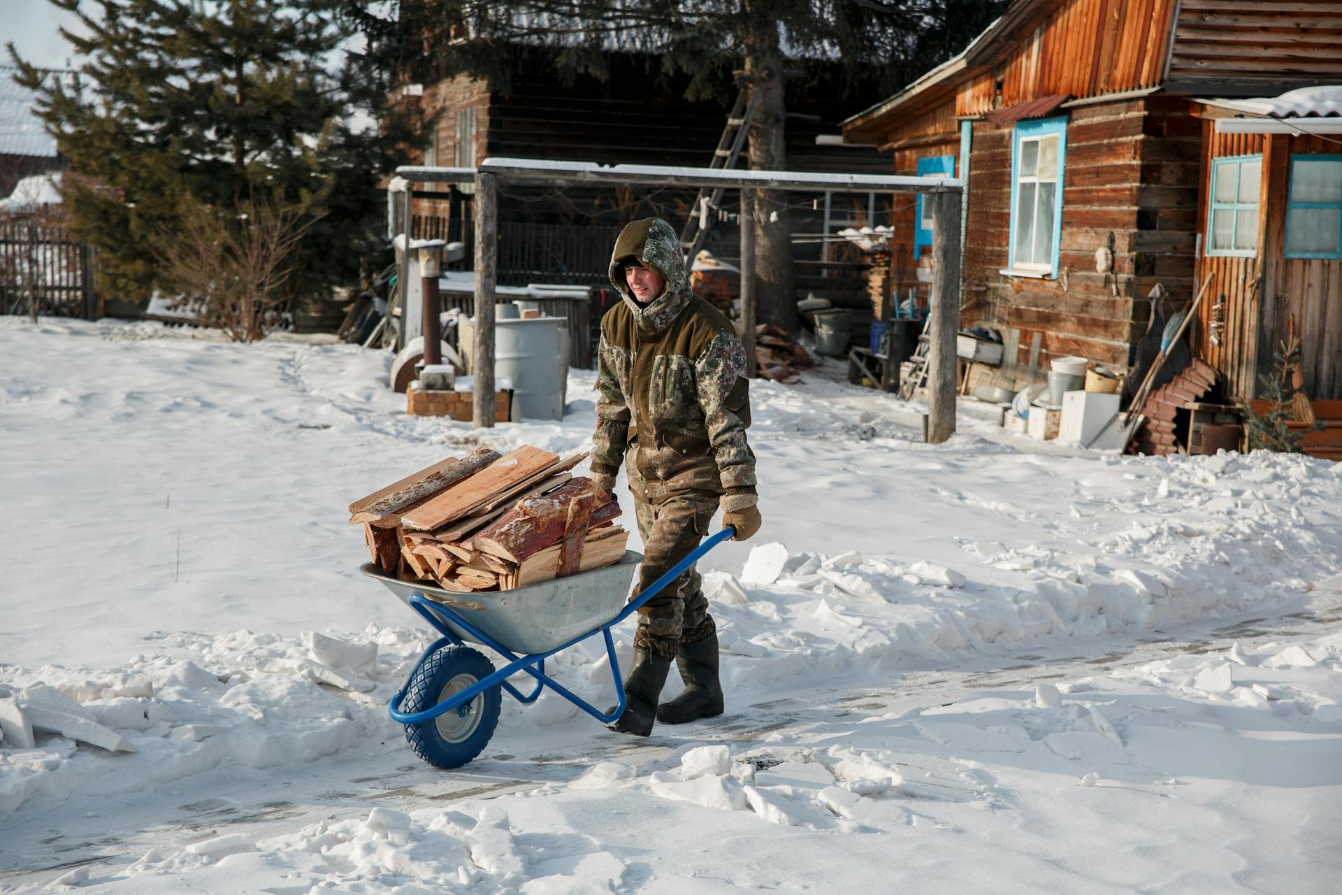 A man in winter gear pushes a wheelbarrow of firewood through a snowy village scene.