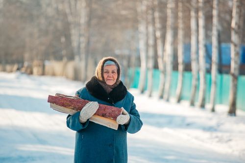 Free Elderly woman in a winter coat holding firewood outdoors on a snowy day. Stock Photo