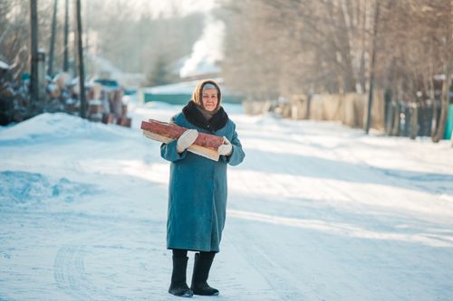Free Elderly woman in winter coat holding firewood on a snowy street. Stock Photo