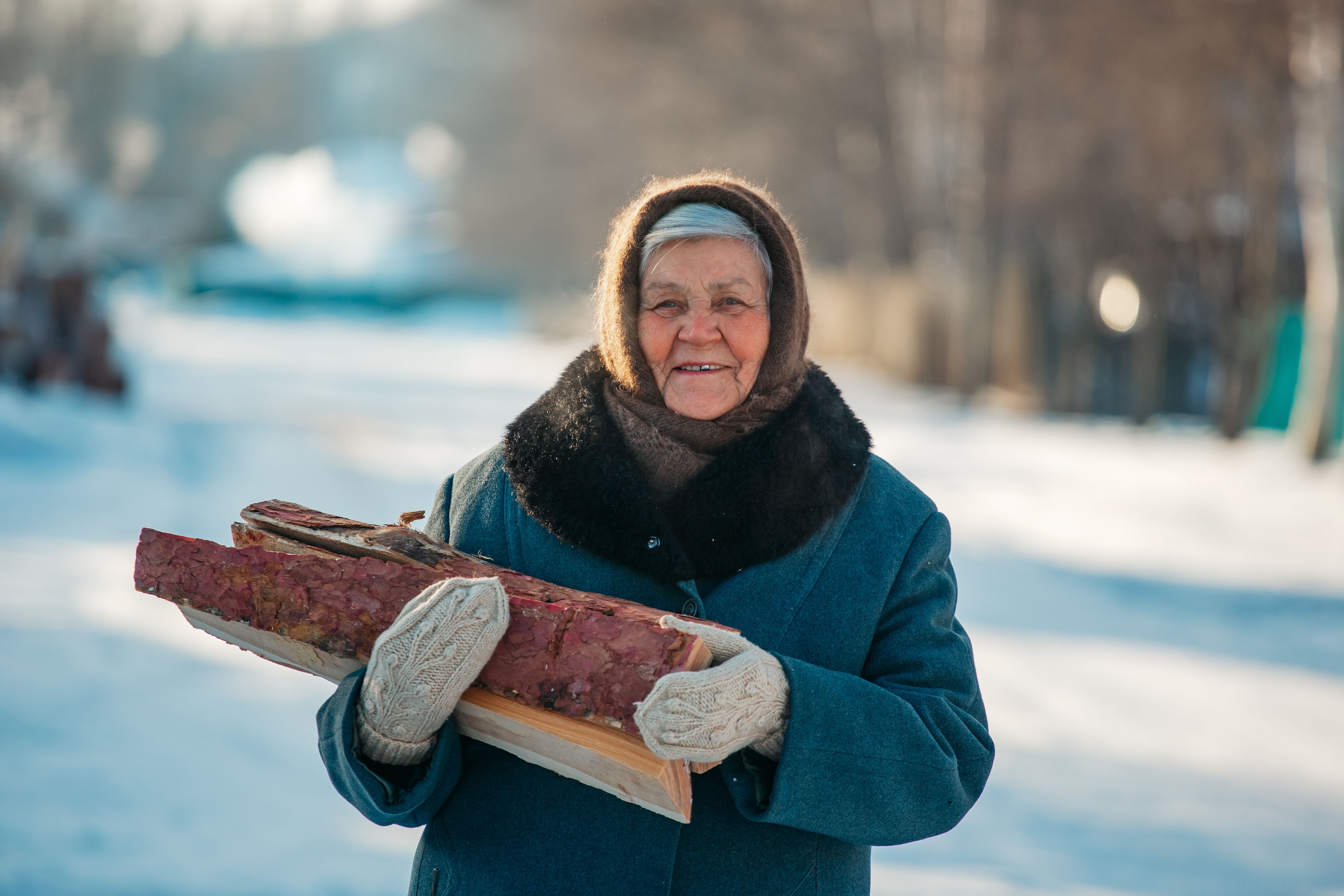 Elderly woman smiling while holding firewood in a snowy outdoor setting.