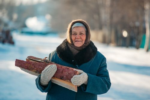 Free Elderly woman smiling while holding firewood in a snowy outdoor setting. Stock Photo