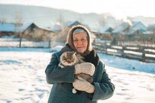 Free Senior woman in winter clothes holding a fluffy cat outdoors in snow covered village. Stock Photo
