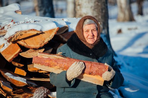 Free Elderly woman holding firewood in snowy winter landscape, wearing warm clothes. Stock Photo