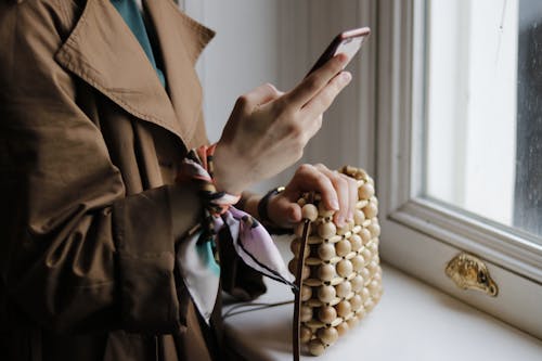 Crop woman with stylish handbag chatting on smartphone against window