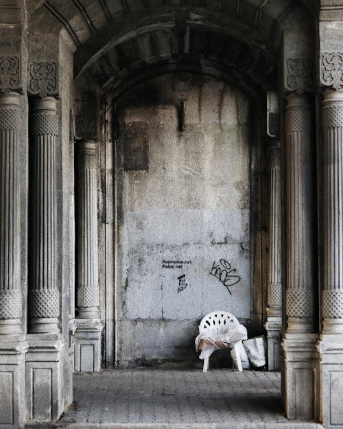 Chair on tiled walkway with aged stone building with ornamental colonnade and rough wall in city