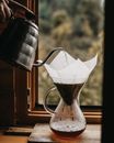 Crop unrecognizable person with kettle pouring hot water into chemex with filter and coffee brewing drink on wooden board on windowsill