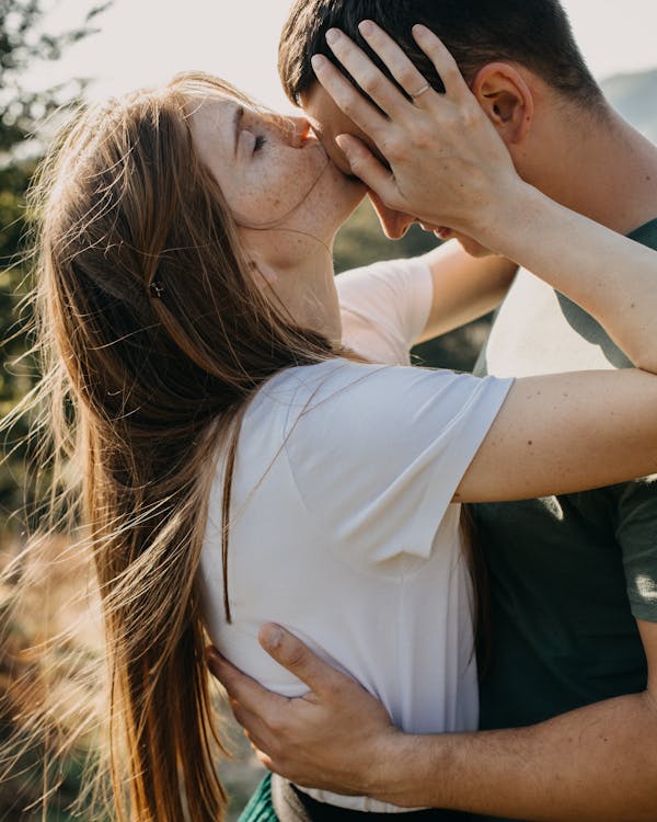 Young romantic couple standing on grassy meadow in countryside embracing while woman kissing man on forehead with tenderness