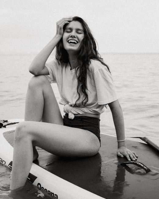 Young happy laughing female wearing bikini and t shirt sitting on sup board with elbow leaning on knee and touching face with hand on water surface