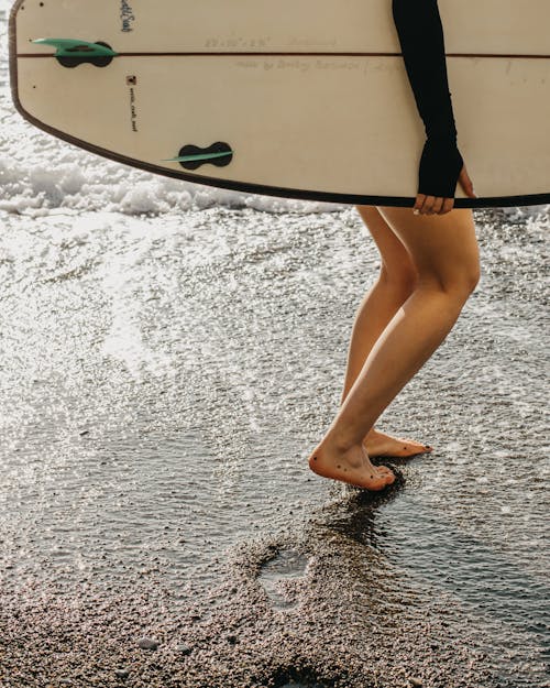 Free Crop side view of anonymous barefoot female surfer holding surfboard in hand while walking along seashore and leaving traces on sunny day Stock Photo