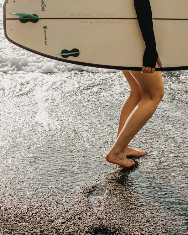 Crop side view of anonymous barefoot female surfer holding surfboard in hand while walking along seashore and leaving traces on sunny day