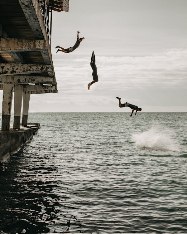 Free Multiple exposure of men in swimwear jumping from pier into calm water of sea in cloudy day Stock Photo