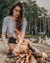Pensive young female with long curly hair in white t shirt sitting on sandy beach near burning bonfire against green trees in nature in daytime