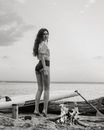 Black and white of pensive female with long curly hair standing on toes on sandy coast with sup board and burning bonfire and looking at camera against calm ocean