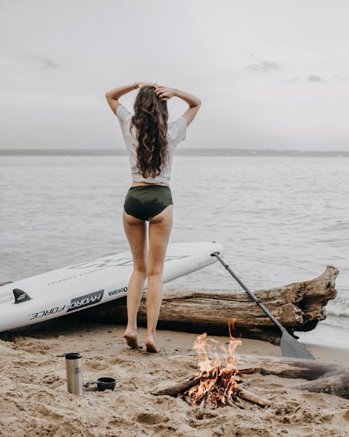 Female standing near sup board on sandy beach with bonfire