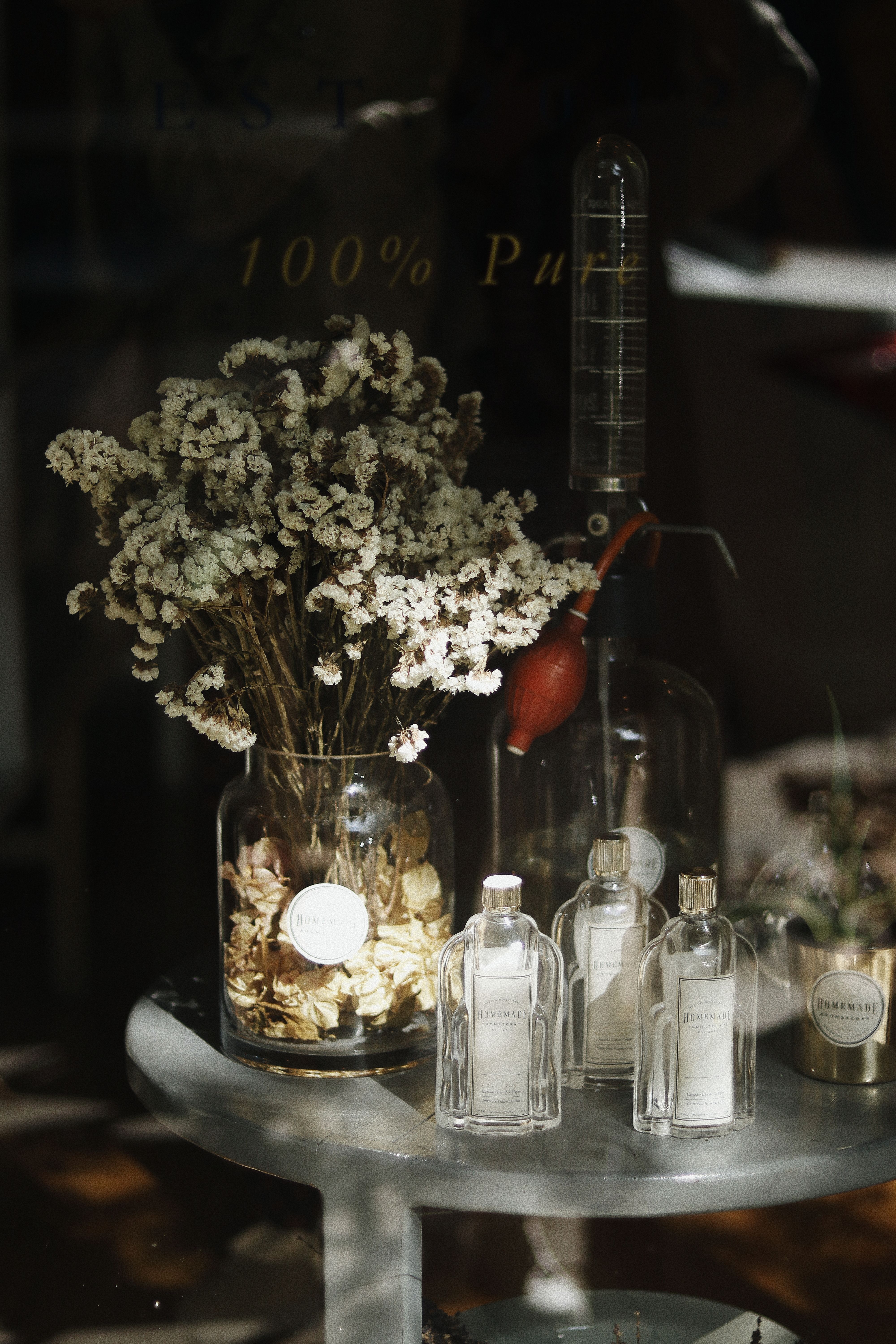 Through glass wall view of blossoming flowers in vase against transparent bottles on table in shop in sun rays
