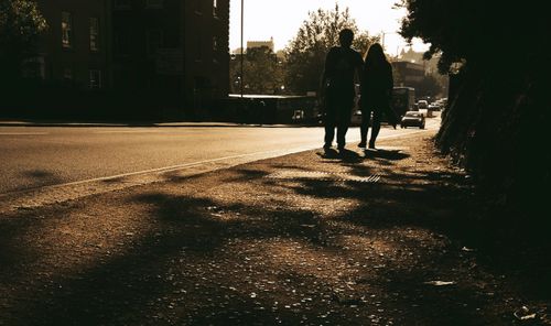Free A couple walks along a city sidewalk at sunset, their silhouettes highlighted against the golden light. Stock Photo