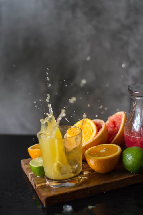 Portion of vitamin juice splashing in glass cup near ripe citruses on cutting board against gray background