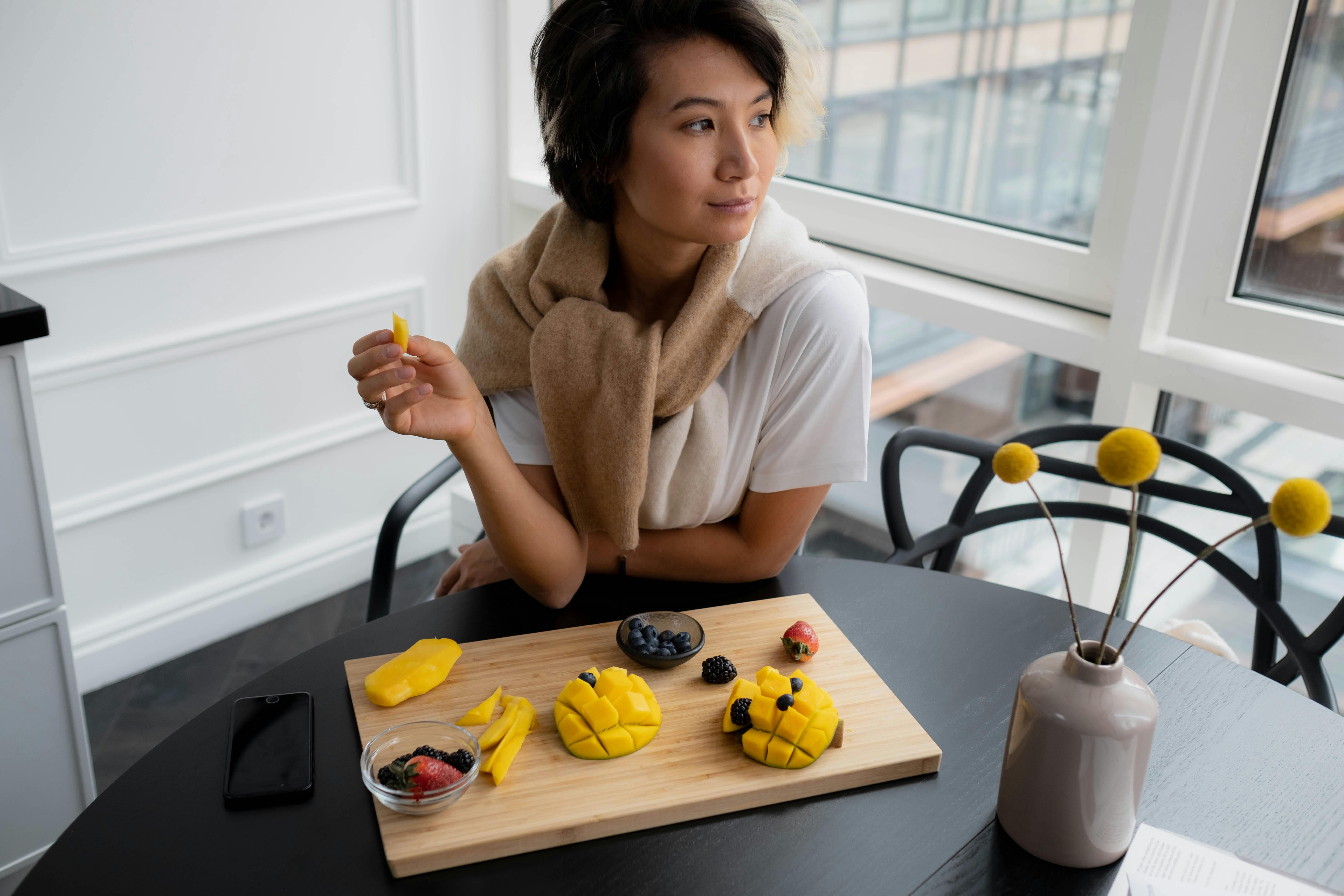 Asian woman savoring sliced mango and berries on a wooden board in a cozy indoor setting.