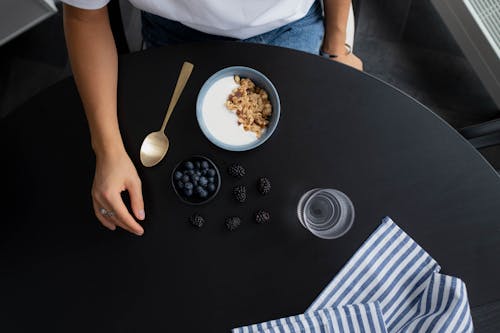 Free Woman in White Shirt Sitting at Black Table With Bowl with Milk and Granola Beside Blueberries and Kitchen Towel Stock Photo