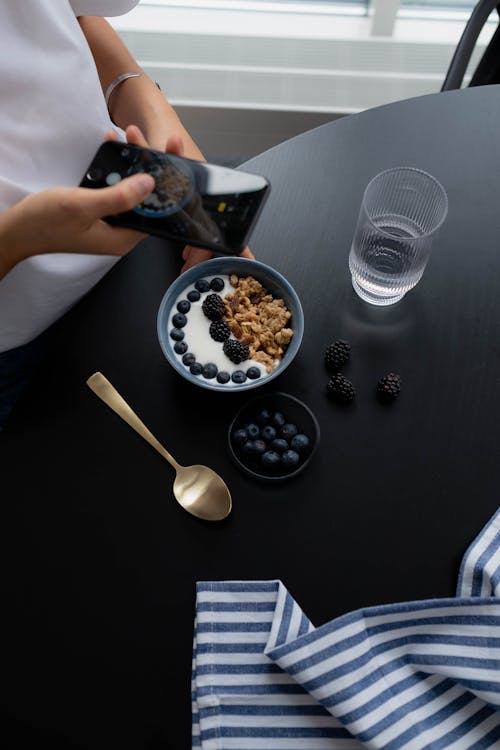 Woman Taking Photo of Granola with Blueberries in a Bowl on Black Table