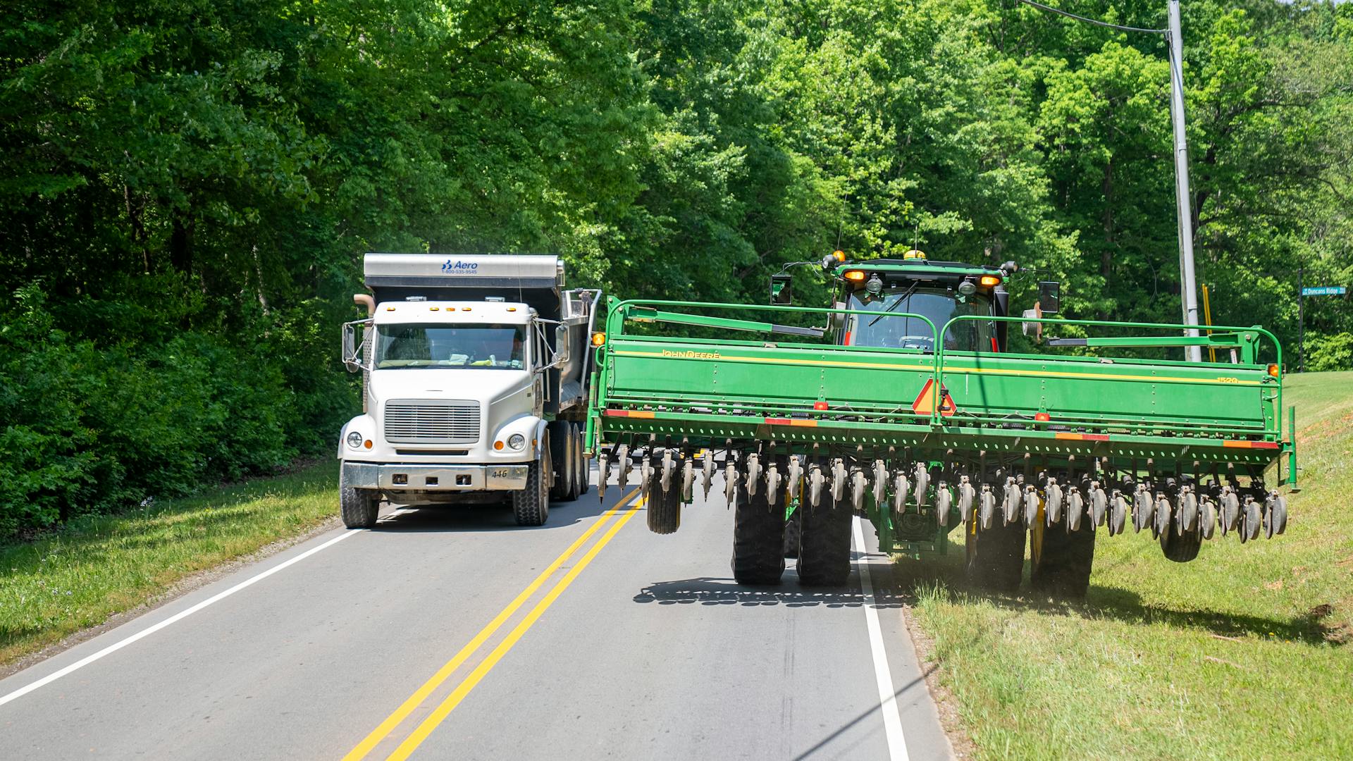 A tractor and dump truck pass on a rural road in North Carolina amidst lush green trees.