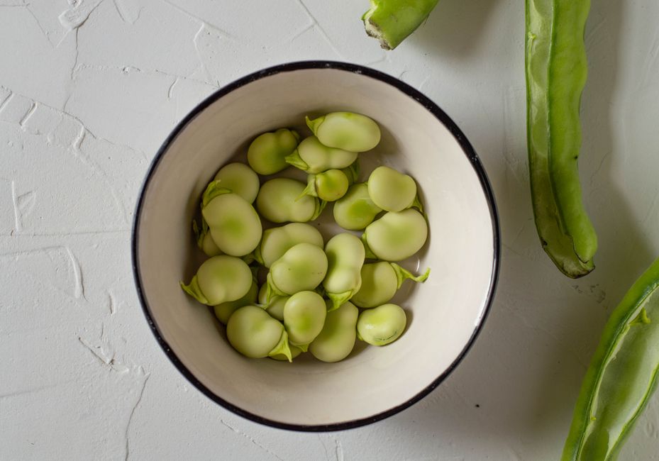 A bowl of bright green shelled fava beans next to a few unopened pods and loose peeled skins on a wooden board
