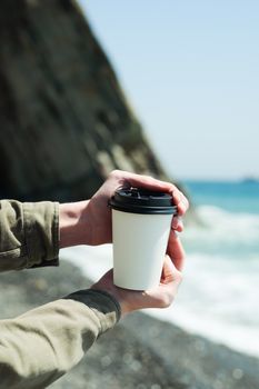 Person holding a disposable coffee cup at the beach with ocean background.