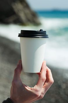 A hand holding a blank paper cup with lid in front of an ocean view, ideal for mockup use.